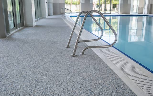Indoor pool deck finished in light grey rubber surfacing beside a pool ladder